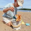 Otrok na plaži z vedrom in igračami za ustvarjanje oblik v pesku, ki so narejene iz okolju prijaznih materialov.