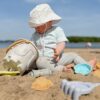 Malček sedi na plaži z vedrom in igračami za pesek, obdan z naravo in peskom.